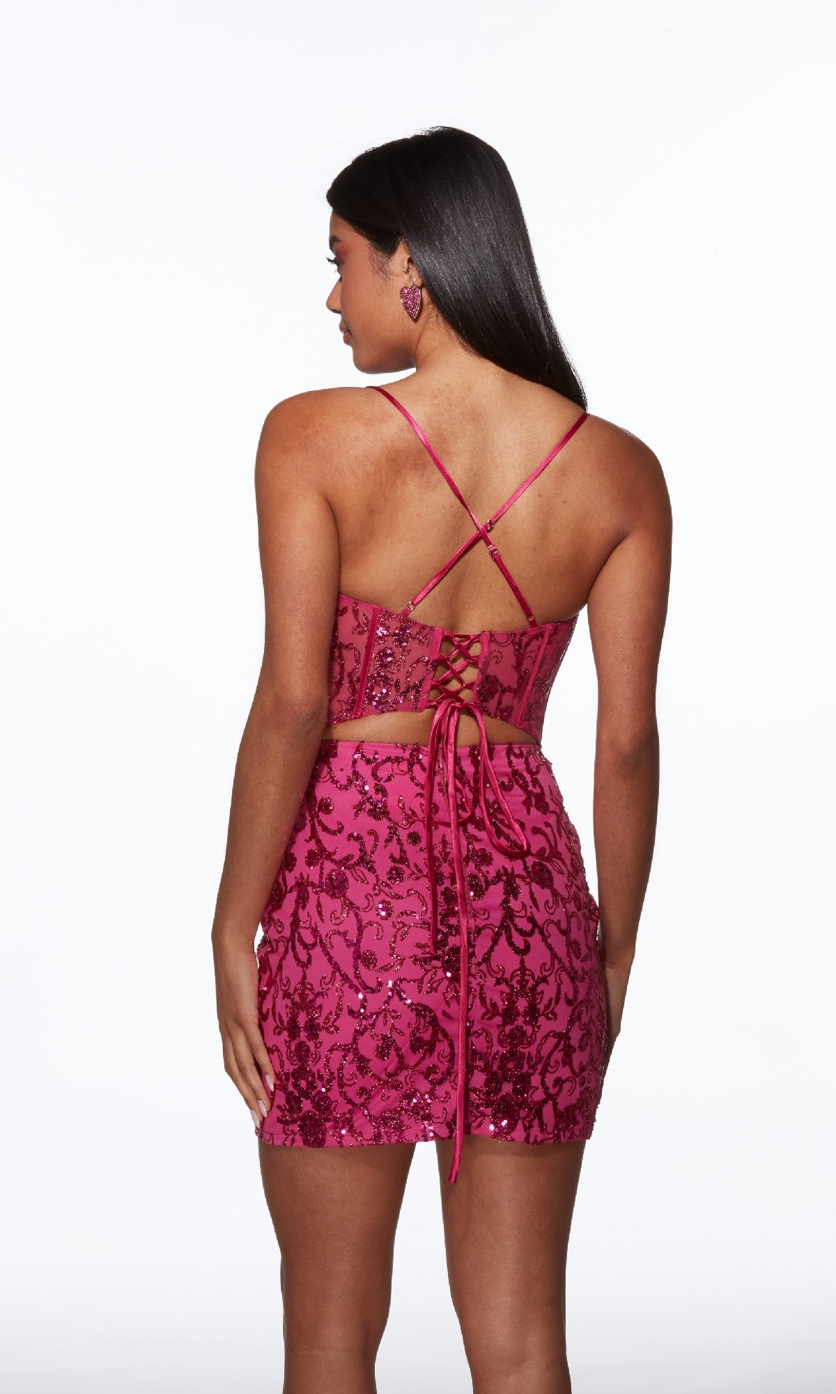 A woman models the Raspberry Short Beaded Hoco Dress (Alyce 40091), featuring pink sequins, a lace-up back, crisscross straps, and matching earrings, standing against a plain white background.